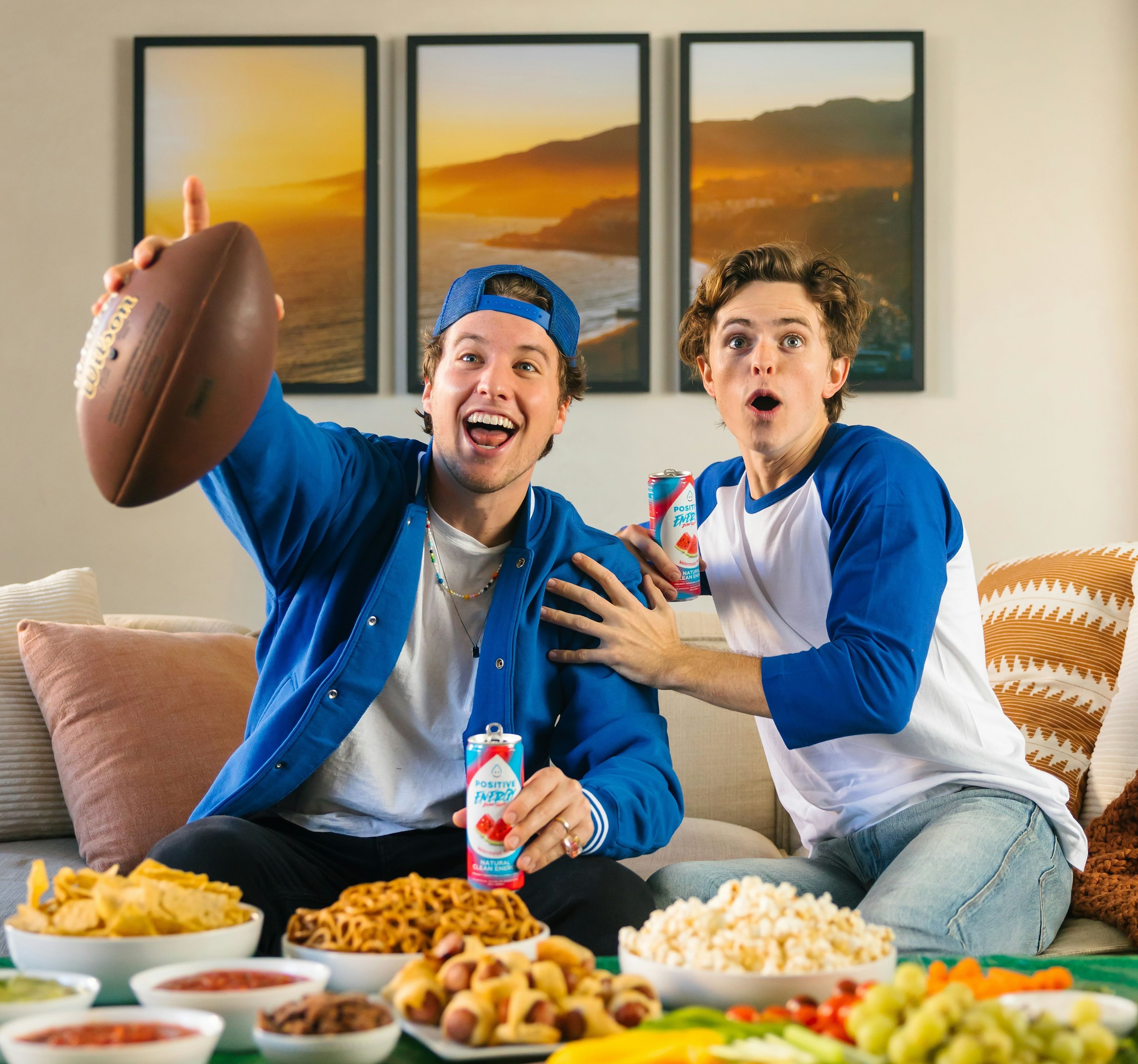 Two people sitting on a couch, pointing excitedly at the tv, with a table full of snacks in front of them.