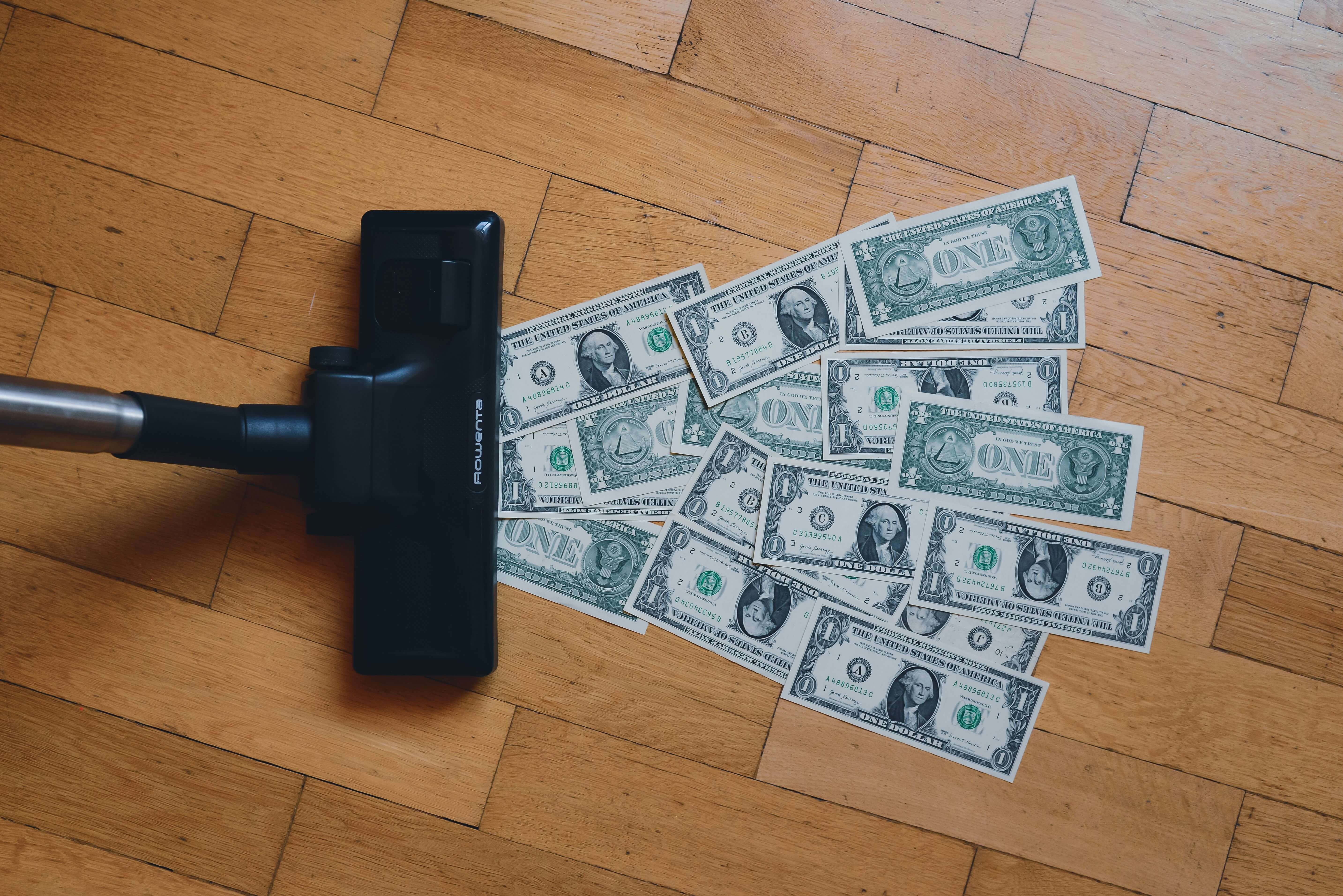 A vacuum sucking up dollar bills off of a wooden floor.